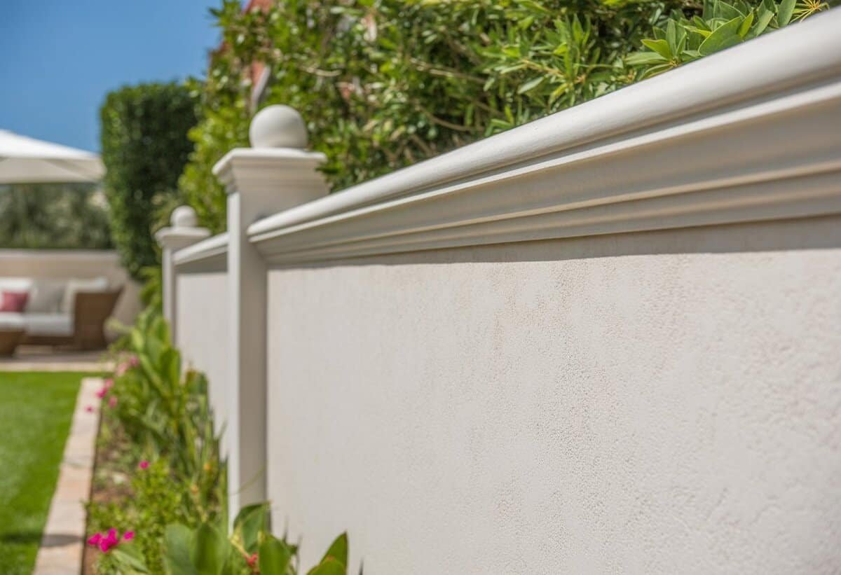 A backyard with a white stucco privacy fence surrounded by green plants and colorful flowers under a clear blue sky.